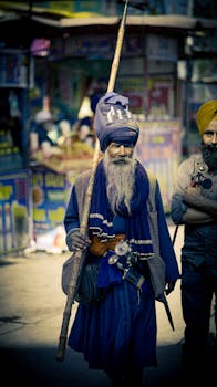 A traditional Sikh elder in blue attire with a staff on a vibrant city street.
