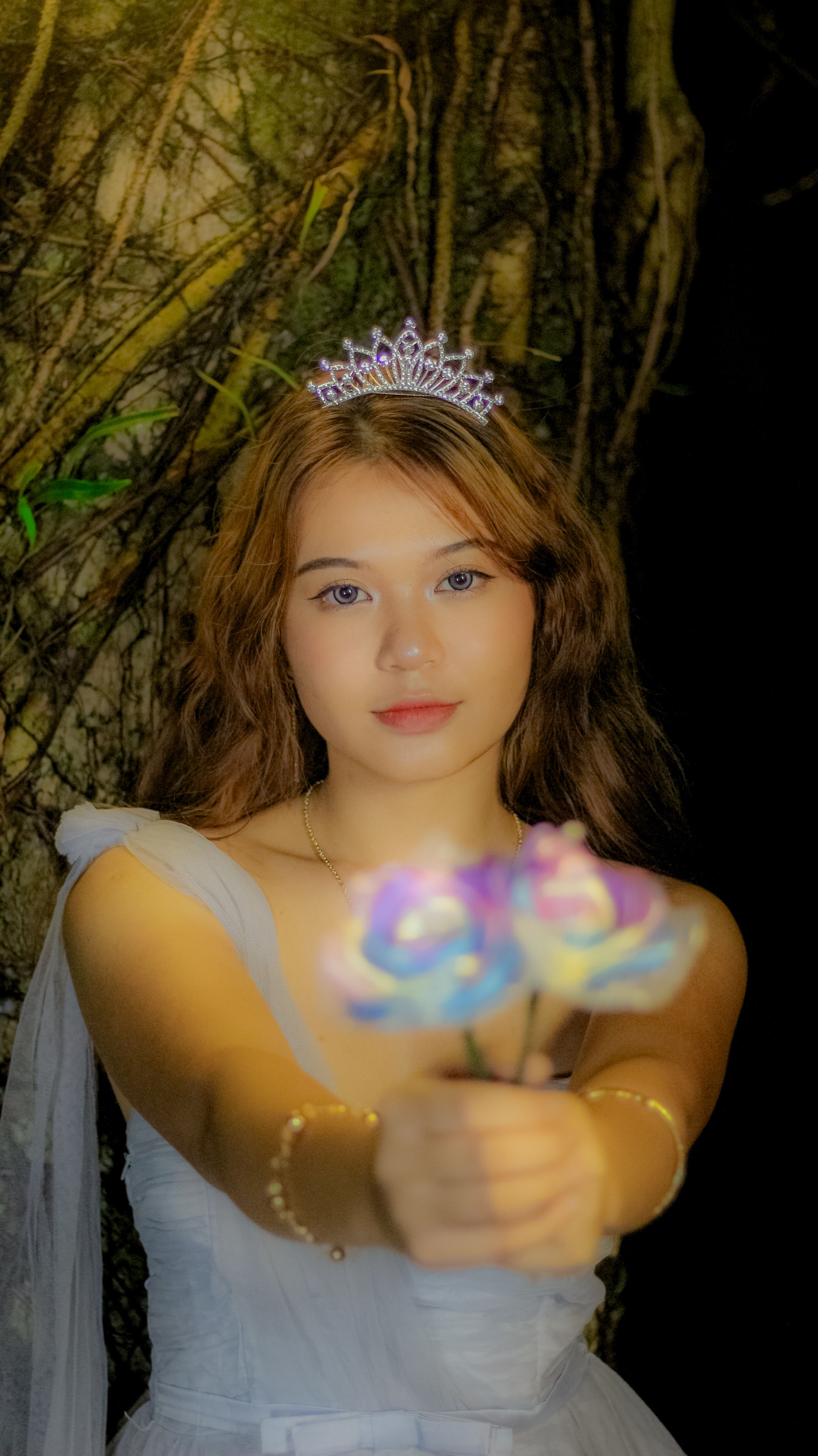 A woman in a white dress with a tiara presents pastel flowers in a magical forest setting.