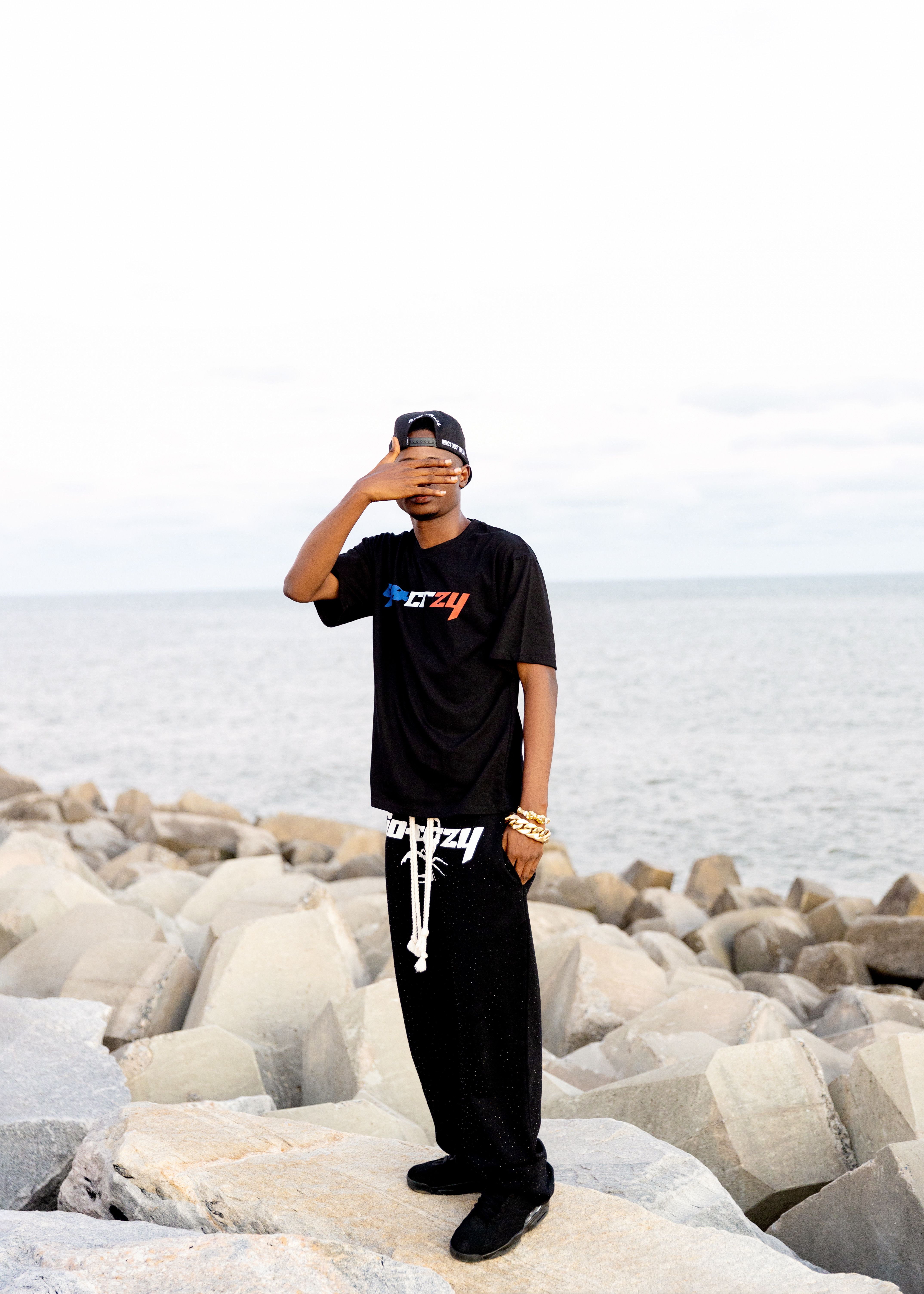A young man in casual clothing poses on a rocky beach with the ocean in the background.