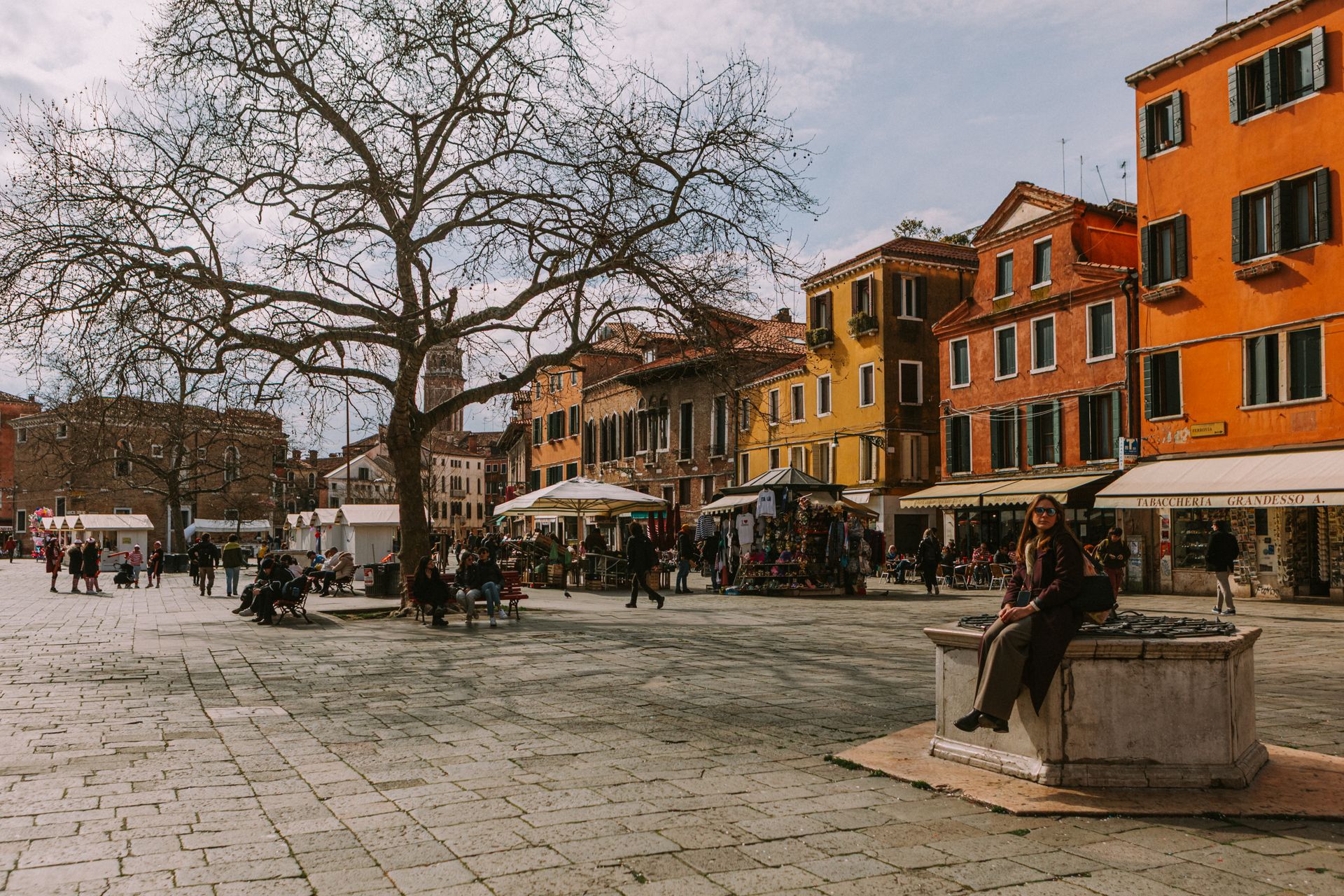 Lively Venetian square with colorful buildings, a leafless tree, and people enjoying the day.