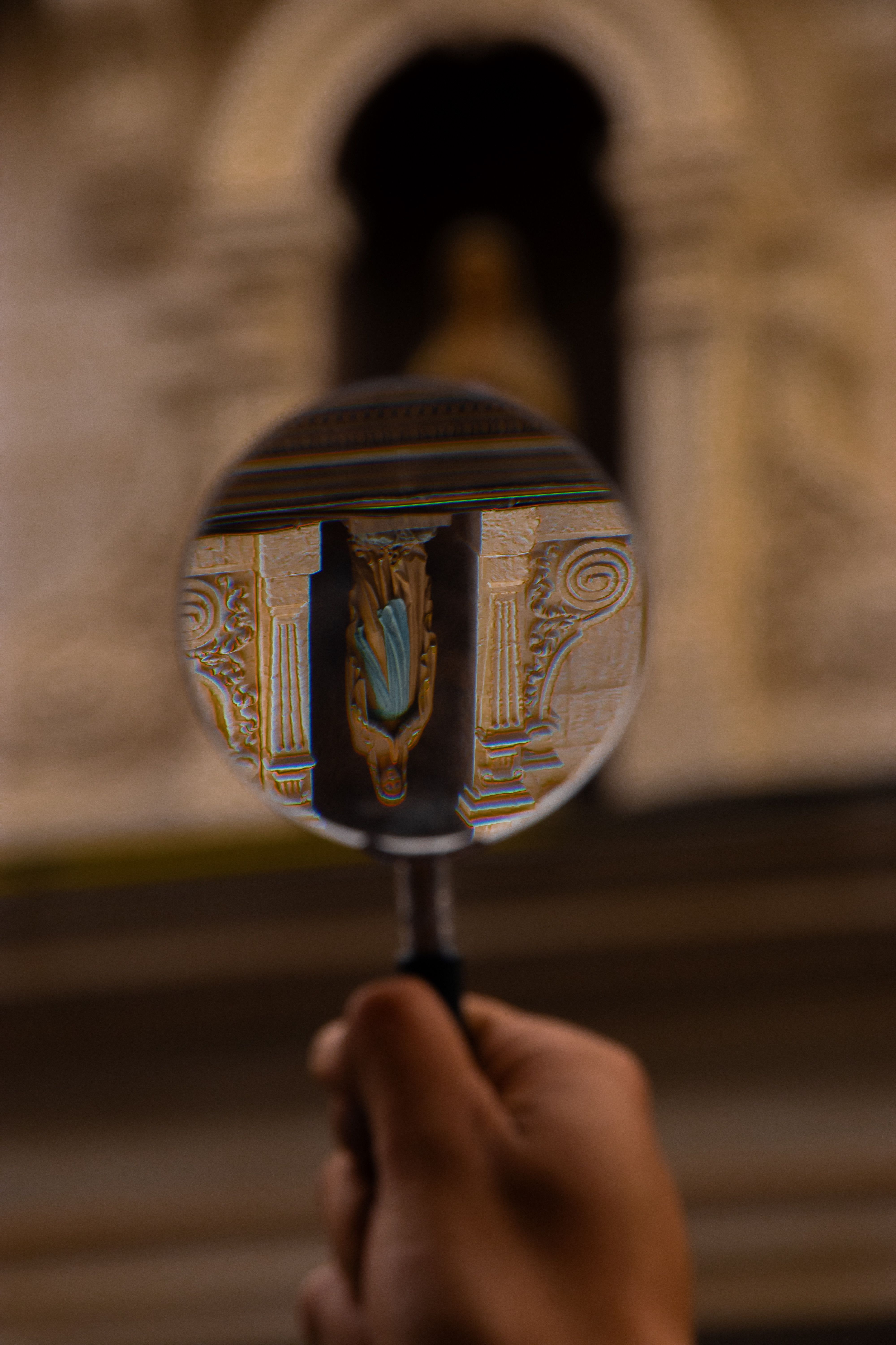 A hand holds a magnifying glass focusing on an ornate statue