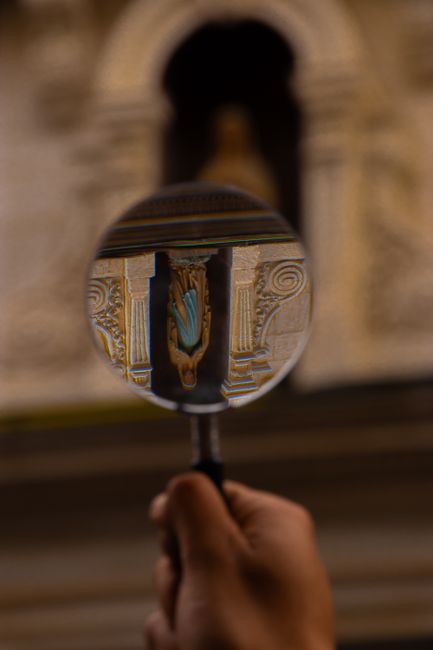 A hand holds a magnifying glass focusing on an ornate statue