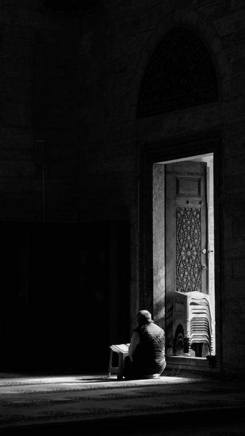 Free Moody black and white image of a person sitting inside, near a door with chairs stacked in shadow. Stock Photo