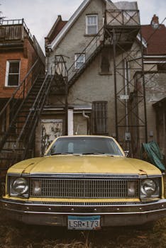 一栋带户外楼梯的旧砖房前，停着一辆经典的黄色汽车