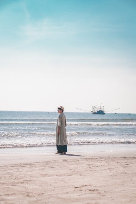 A woman walks along a calm beach with a fishing boat in the distance on a sunny day.