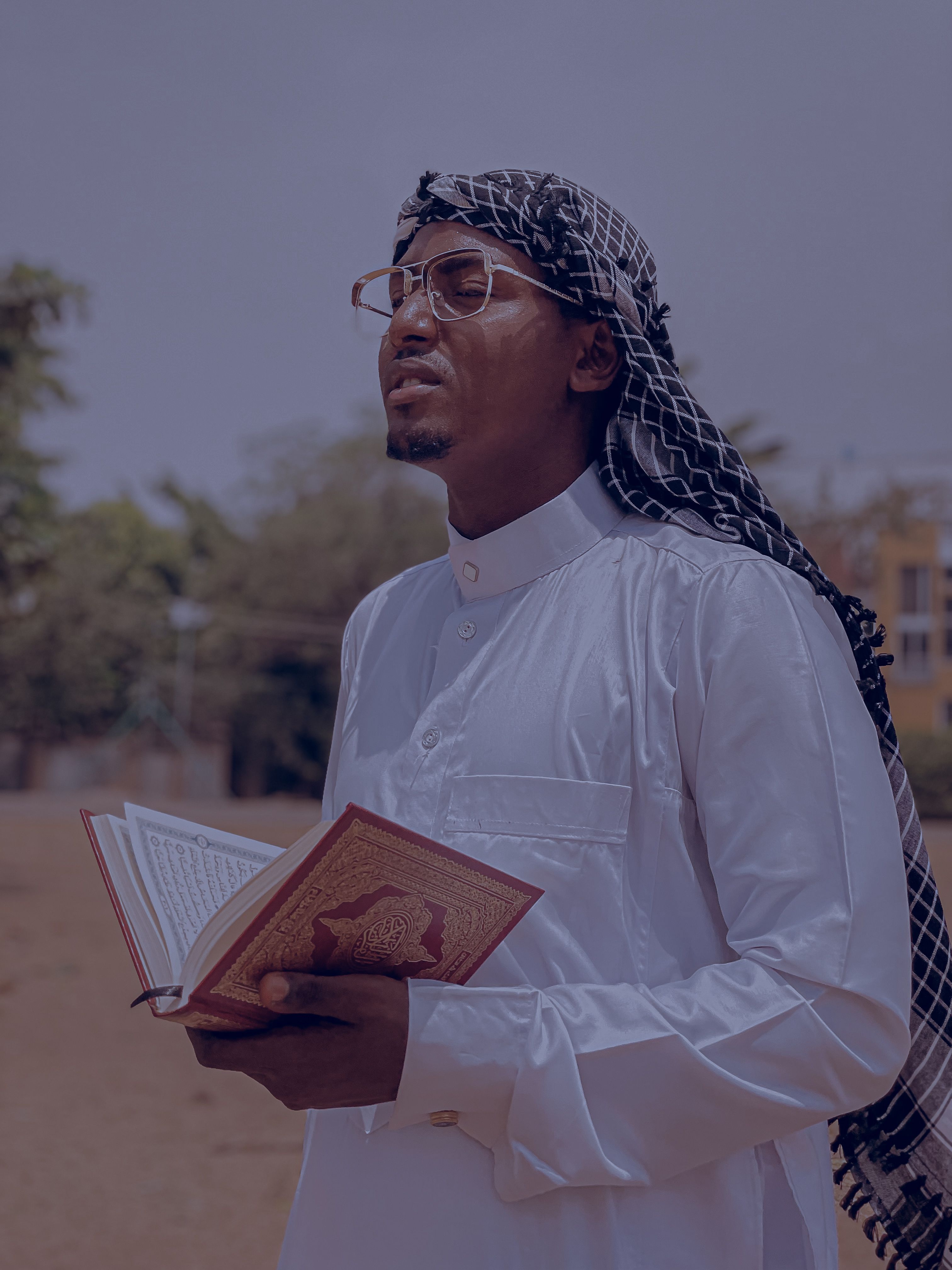 A man in traditional clothing reading a Quran outside on a sunny day.