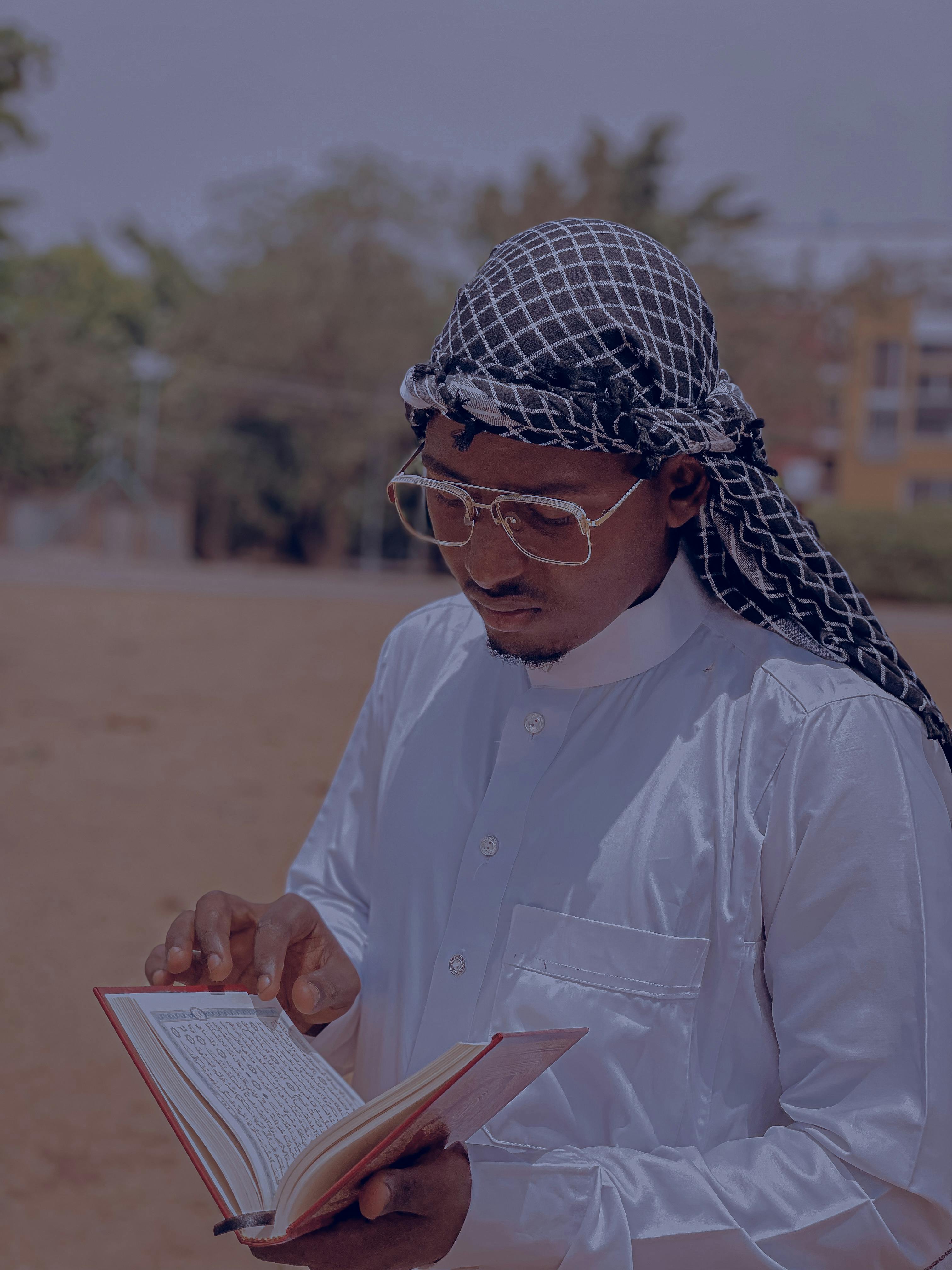 Adult man wearing traditional clothing reading a book outdoors on a sunny day.