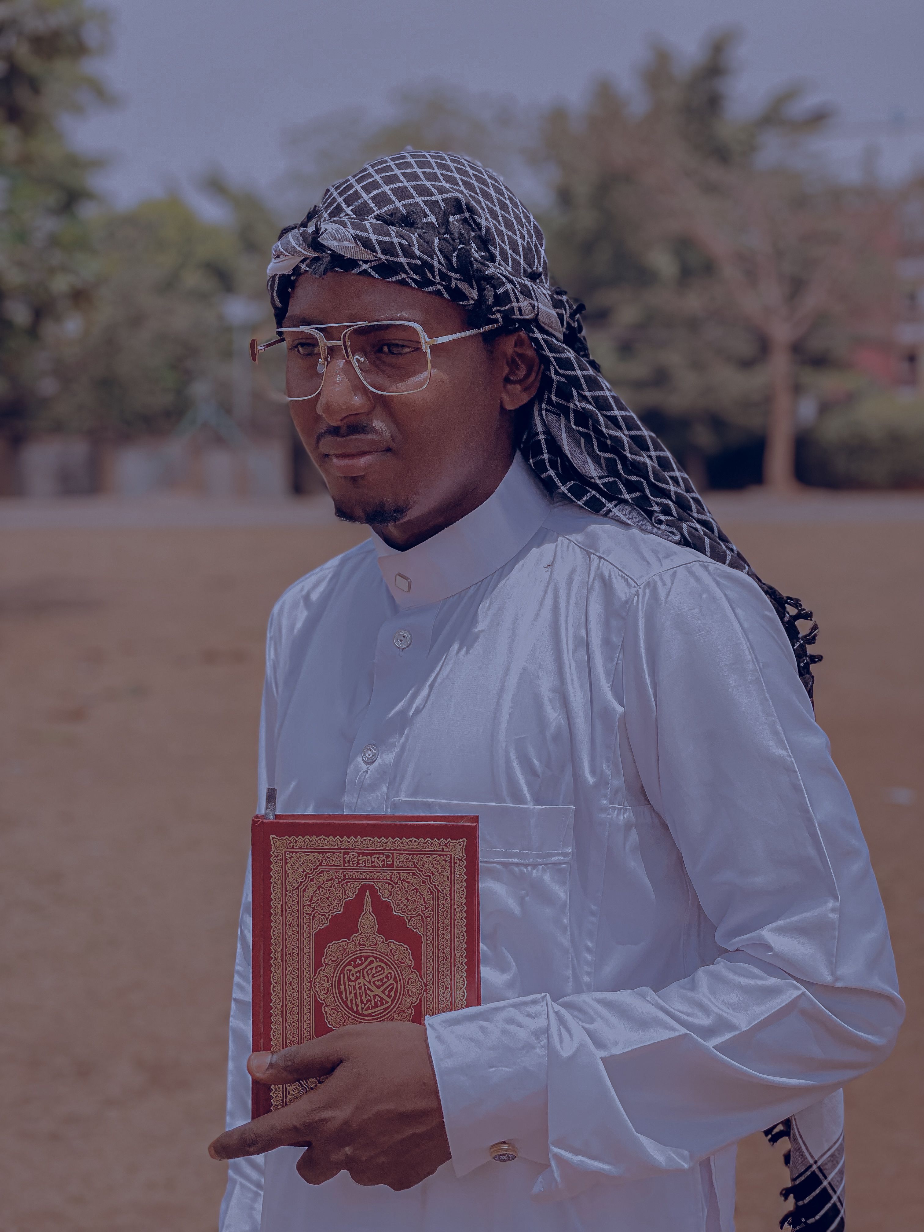 A man in traditional attire holds a Quran in a serene outdoor setting.