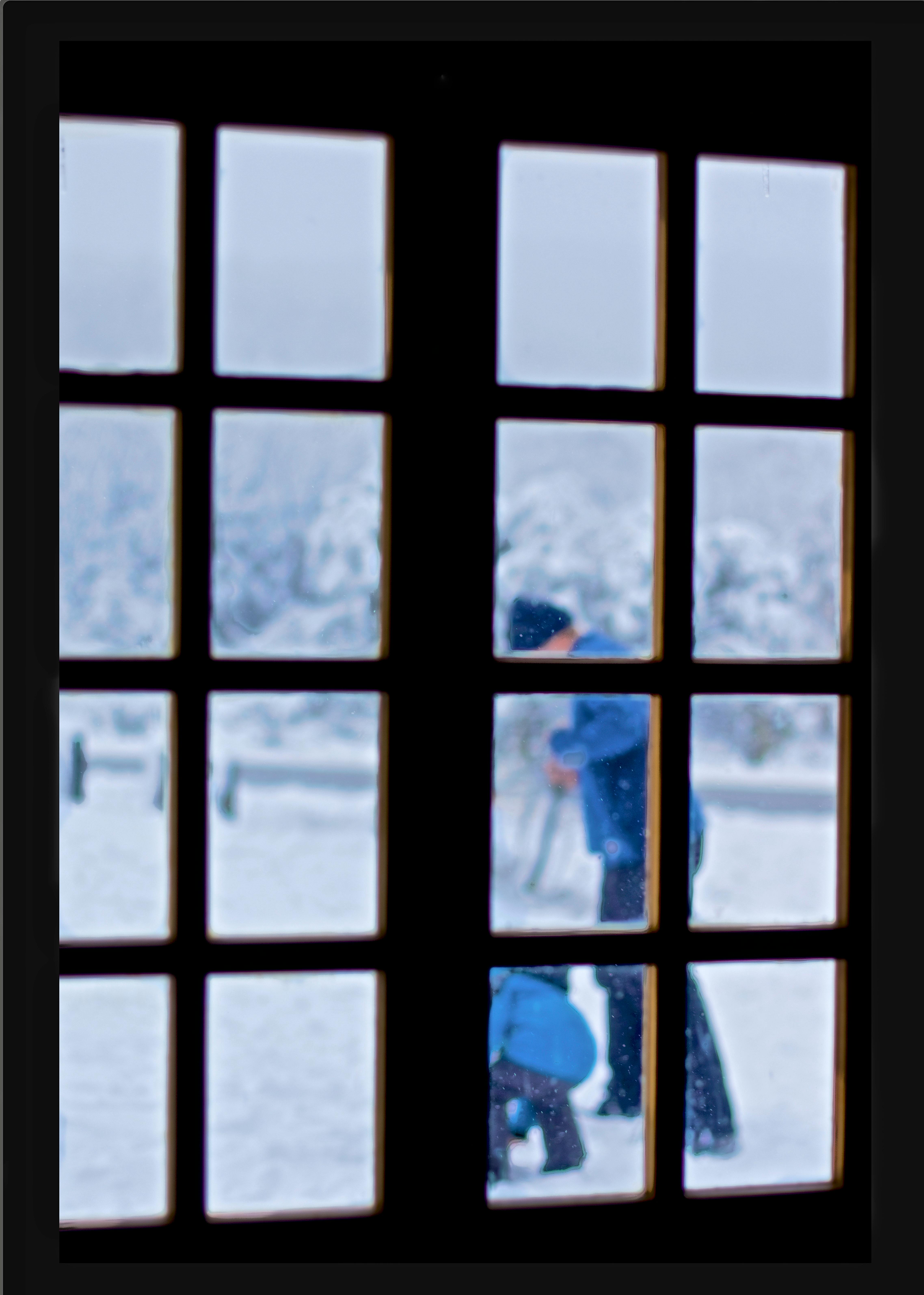 A frosty window frames a snowy scene with blurred figures outside.