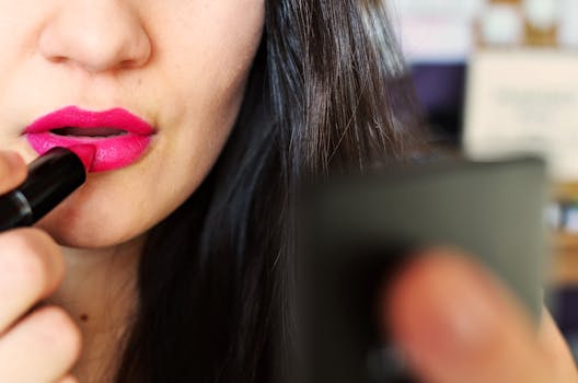 Close-up of a woman applying bright pink lipstick while looking in a mirror, highlighting beauty and makeup.