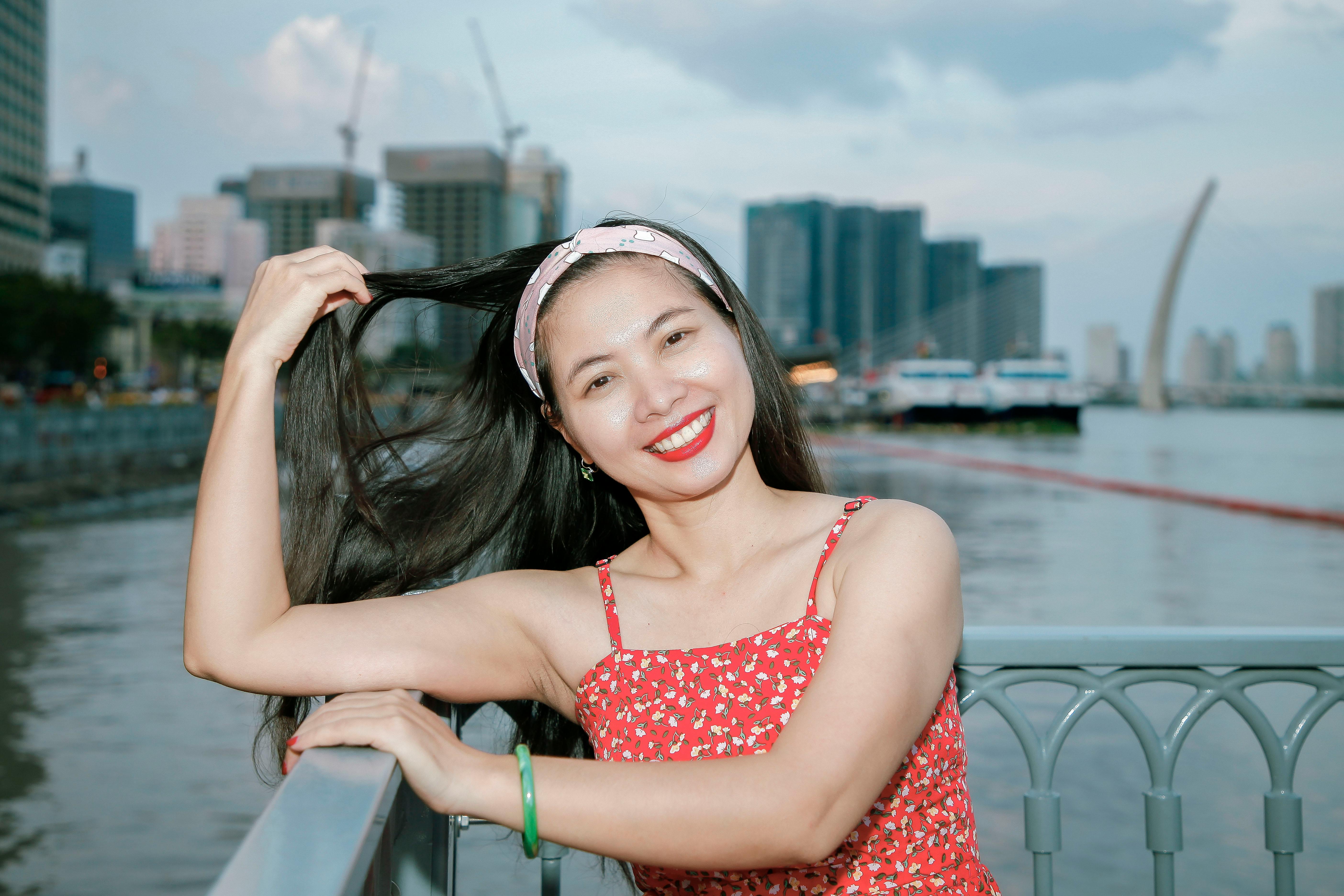Cheerful woman in red dress poses by the river with cityscape in the background.