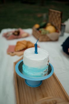 A birthday cake with blue icing and candle on a picnic setup outdoors.
