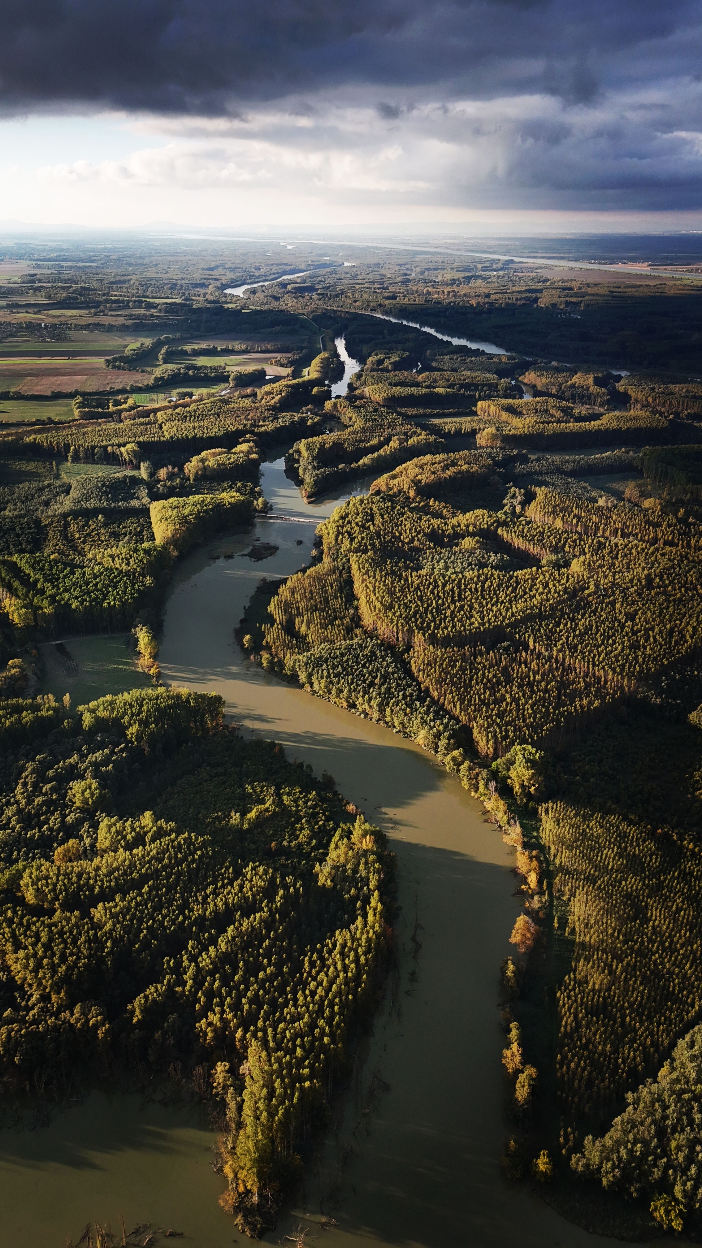 Aerial shot of a winding river surrounded by lush green forests and fields under a dramatic sky.