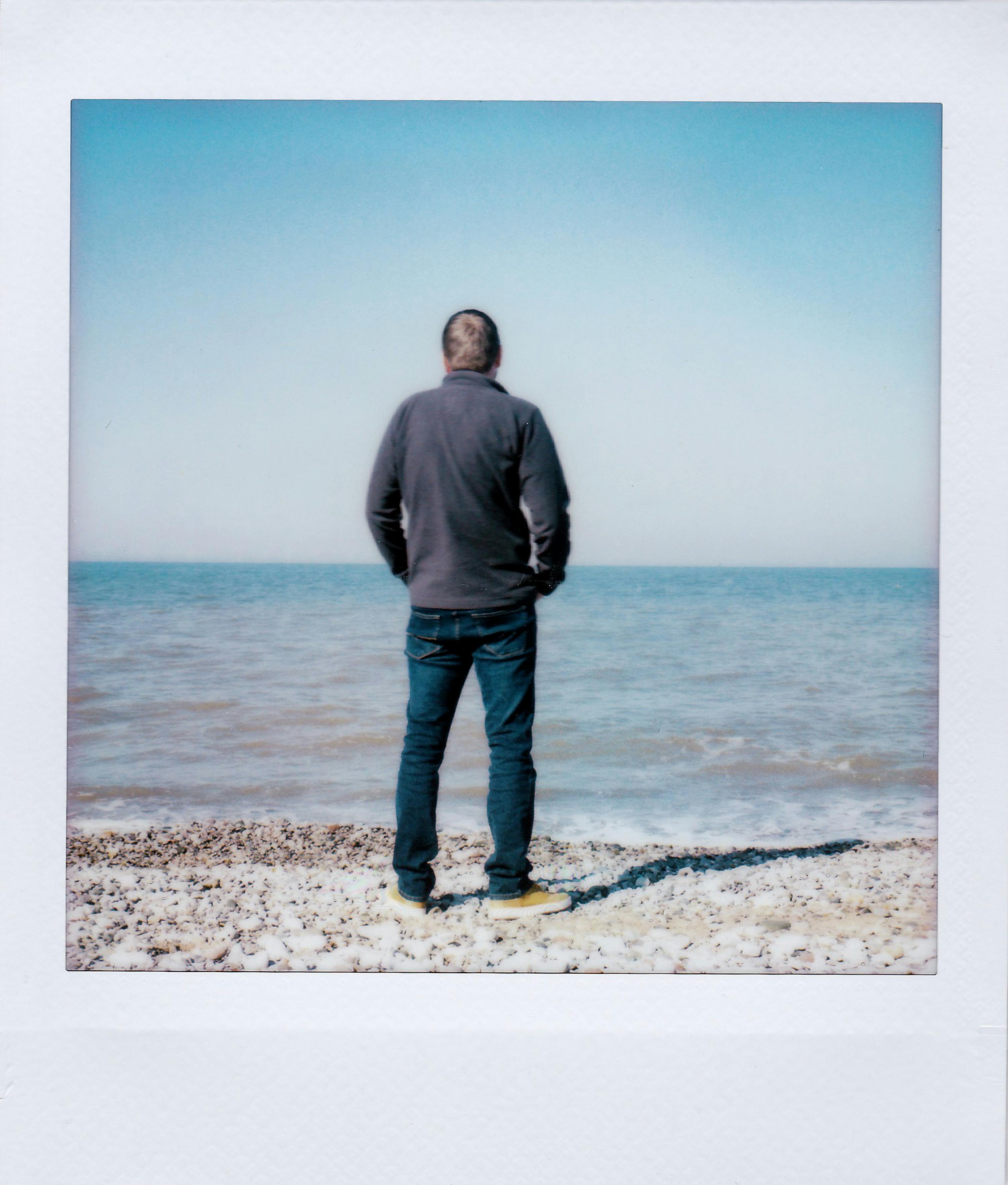 A person stands on a pebble beach facing the sea, evoking solitude and introspection.