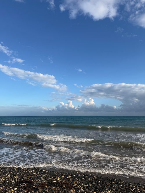 Free Tranquil scene of waves crashing on a pebble beach under a vast blue sky with fluffy clouds. Stock Photo
