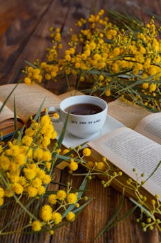 Cozy scene with yellow mimosa flowers, open book, and coffee cup on wooden table.