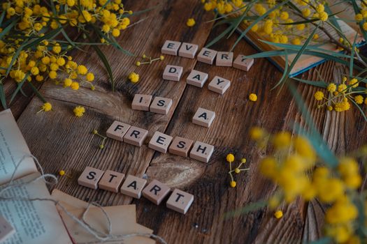 Wooden tiles spell 'Every day is a fresh start' surrounded by yellow flowers on rustic table.