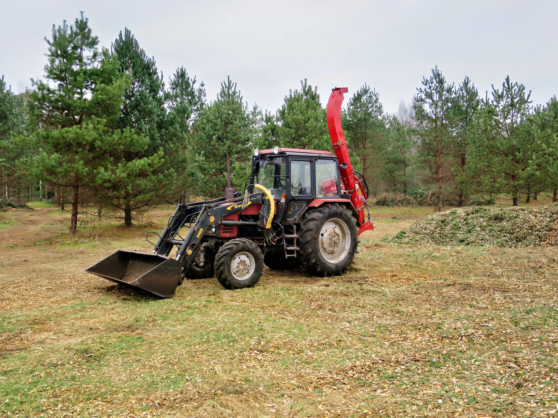 A red tractor equipped for logging and wood chipping, set in a dense forest.