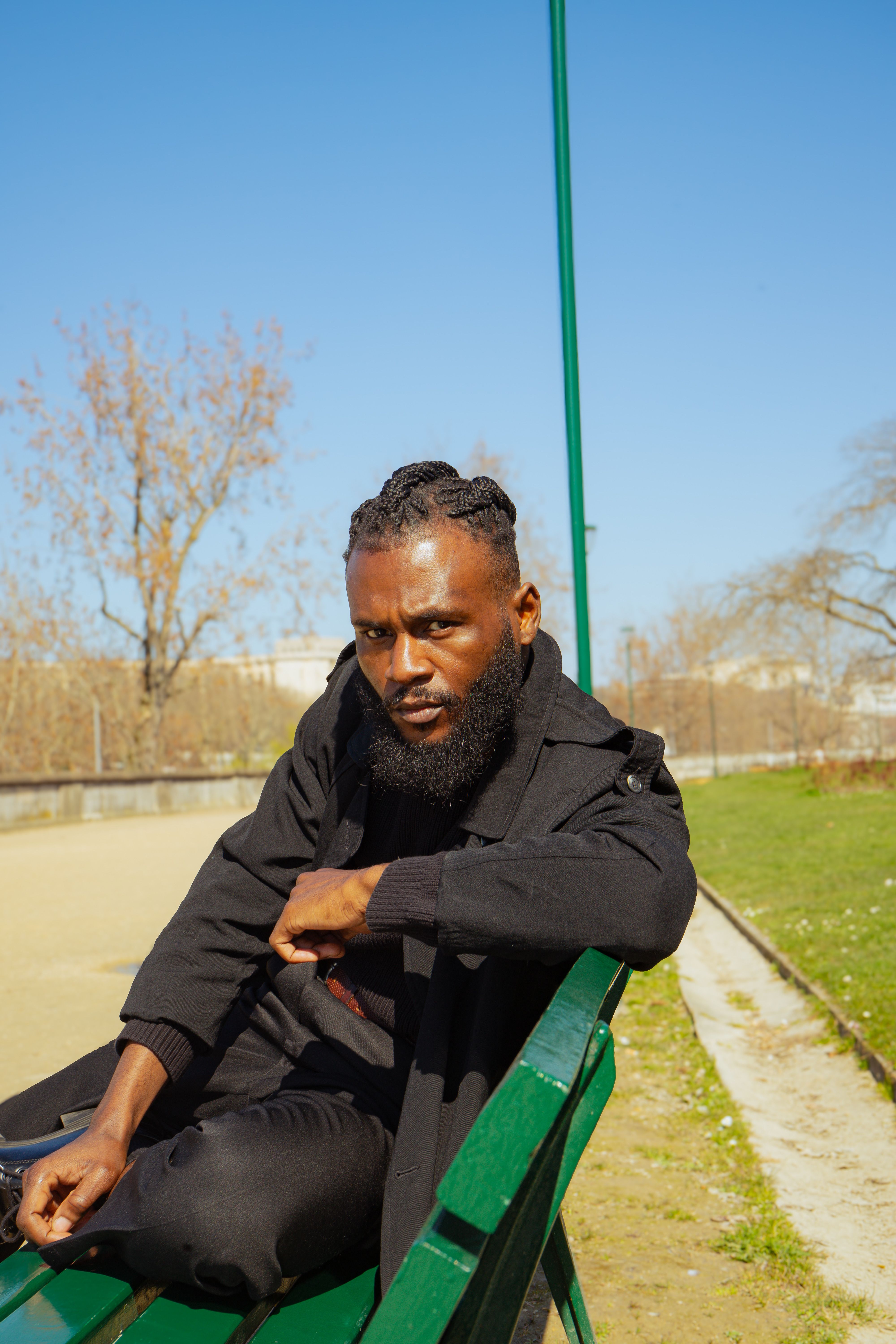Young man with beard sitting on a park bench under clear blue sky.