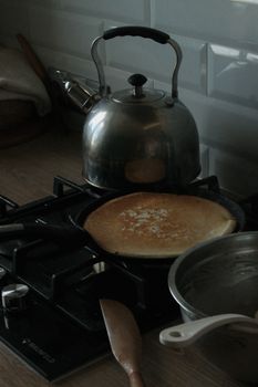 Dark kitchen scene with pancake cooking on a stove and a kettle beside it, creating a homely vibe.