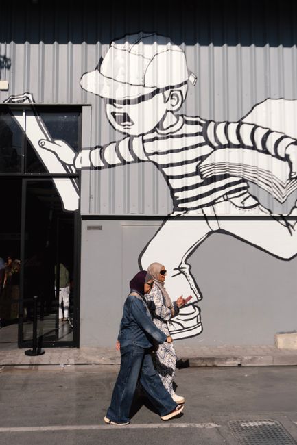 Two women walk past a striking mural on a city street in Dubai.