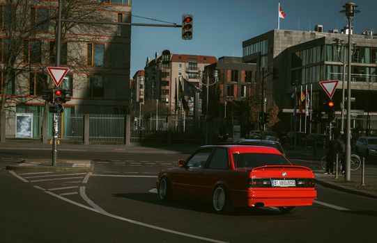 A classic red car waits at a city intersection under bright daylight, surrounded by urban architecture.