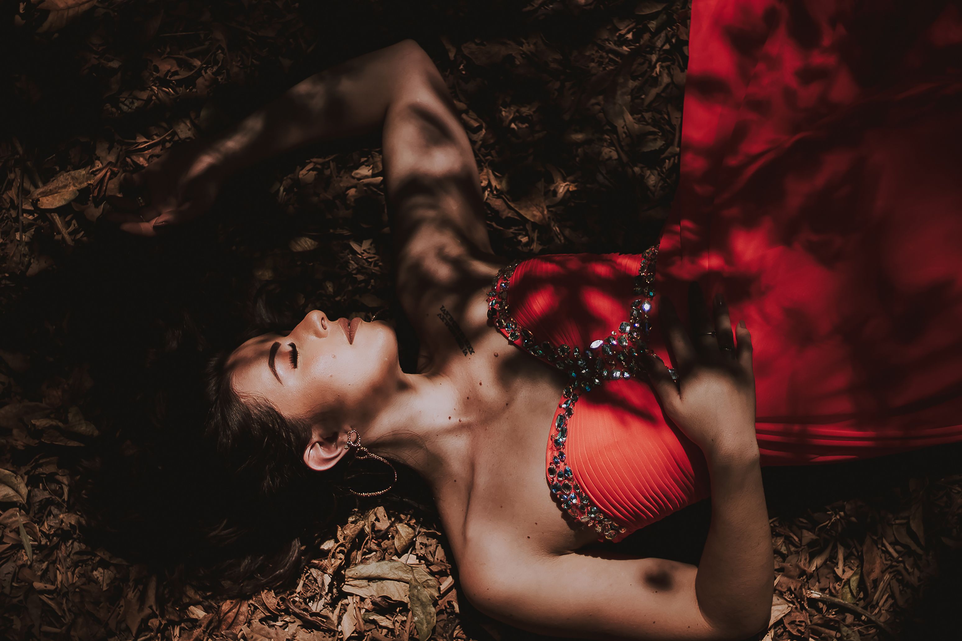 A woman in a red gown rests among fallen leaves, casting a dramatic shadow and showcasing elegant fashion.