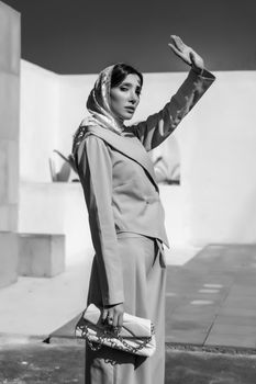Stylish woman in headscarf and suit poses in bright outdoor setting.