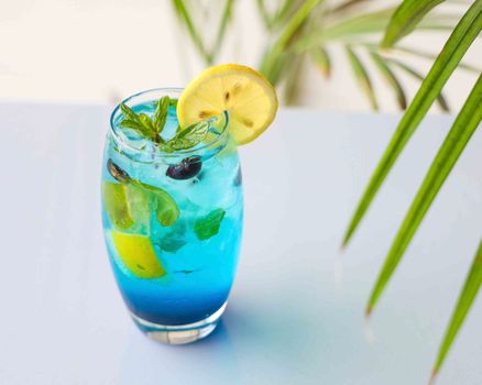 Refreshing blue mocktail with lemon slice, mint, and blueberries on a tropical background.