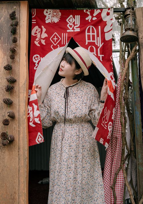 Free Woman in floral dress and hat standing by red tapestry with symbols. Stock Photo