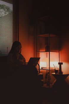 A woman working on a laptop in a dimly lit room with orange ambient lighting and cozy atmosphere.