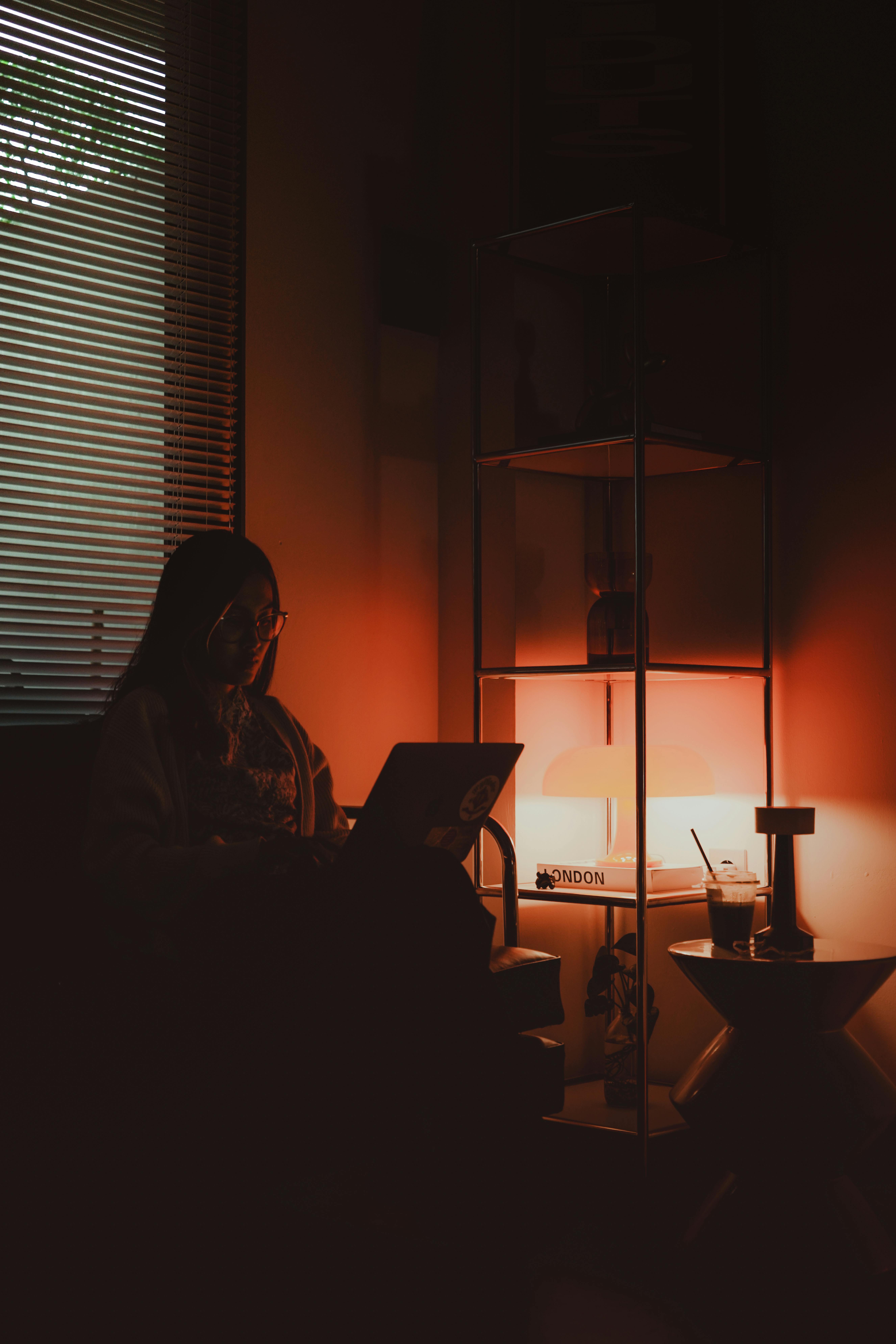 Gratis Seorang wanita bekerja di depan laptop di ruangan remang-remang dengan pencahayaan oranye dan suasana yang nyaman. Foto Stok