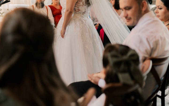 Bride in white dress speaks to seated guests at elegant indoor wedding venue.