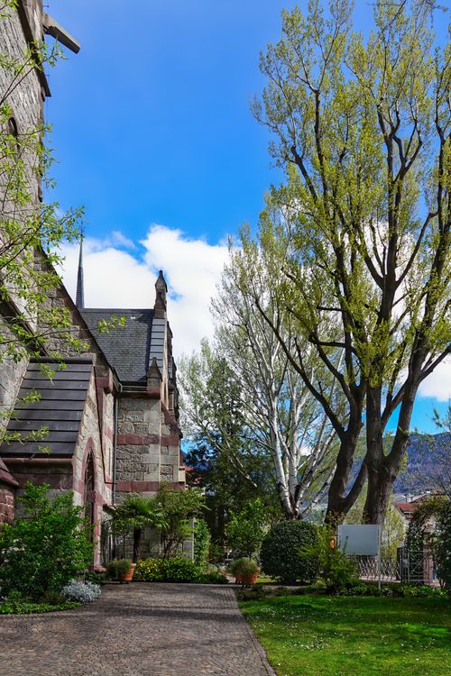 Free A scenic view of a historic church surrounded by lush greenery and a bright blue sky in Meran, Italy. Stock Photo
