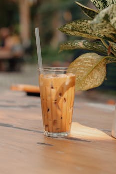 Refreshing iced coffee with straw on a wooden table next to a plant in sunlight.