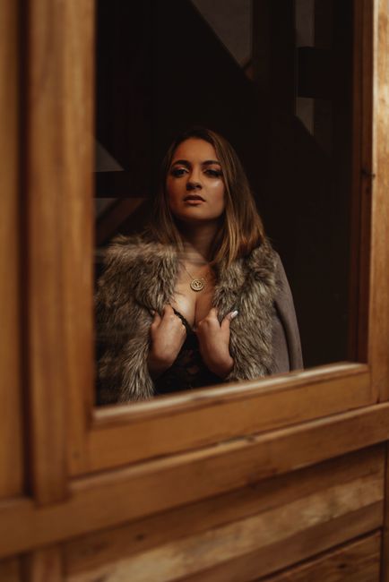 Glamorous woman in fur coat posing elegantly indoors near a wooden window frame.