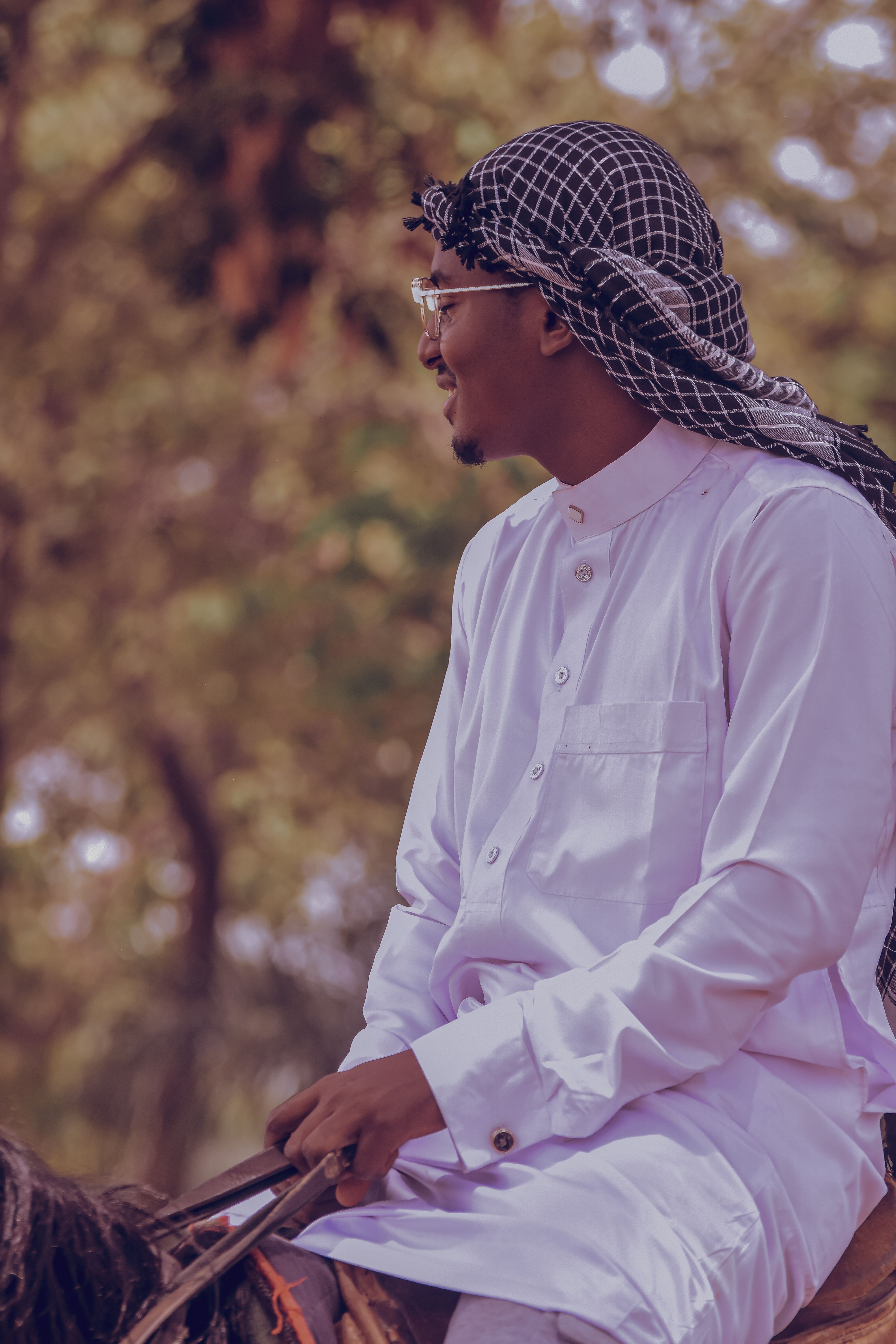 A man wearing traditional attire sits on a horse outdoors, surrounded by trees.