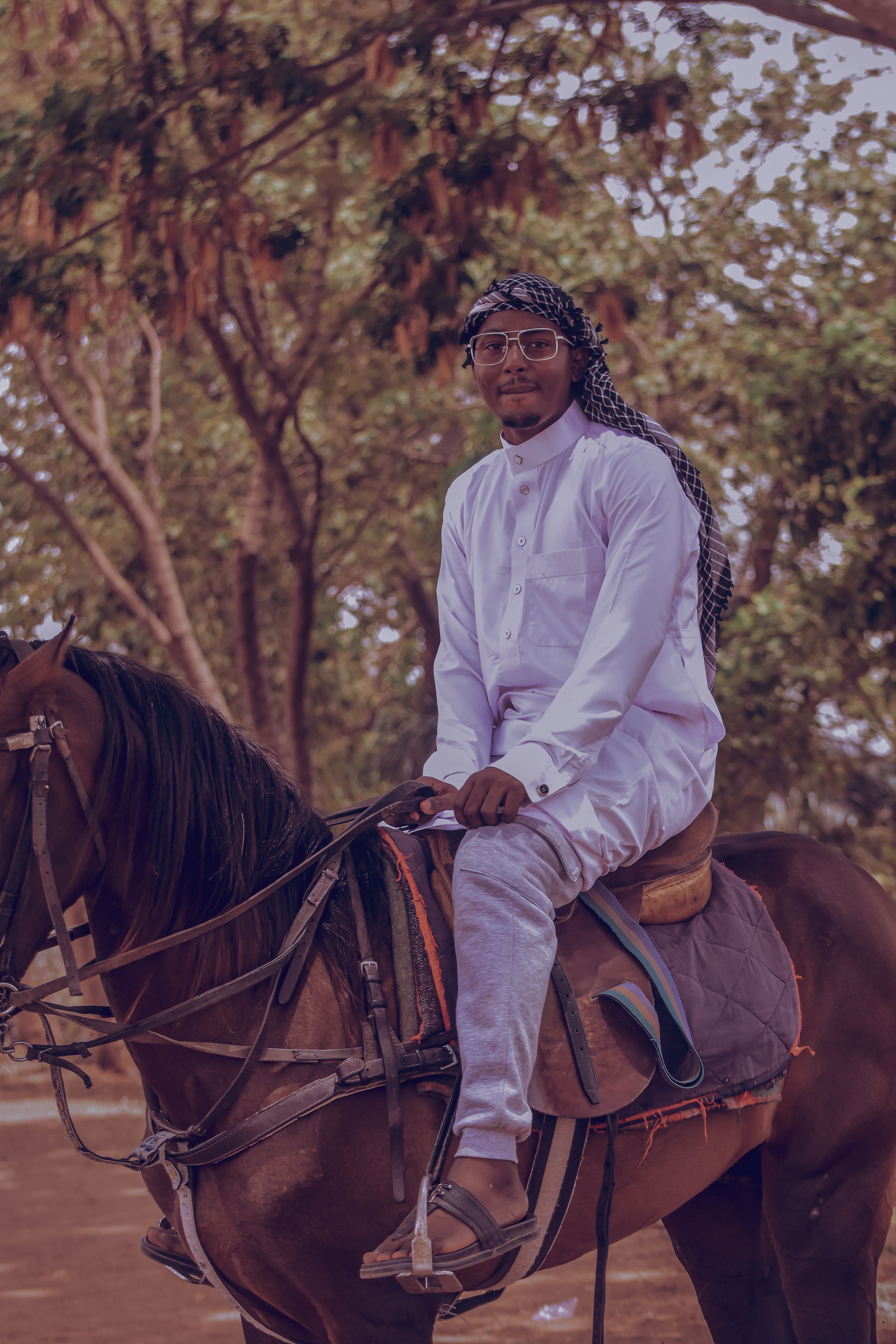 Adult male in traditional attire riding a horse, surrounded by trees in a natural setting.