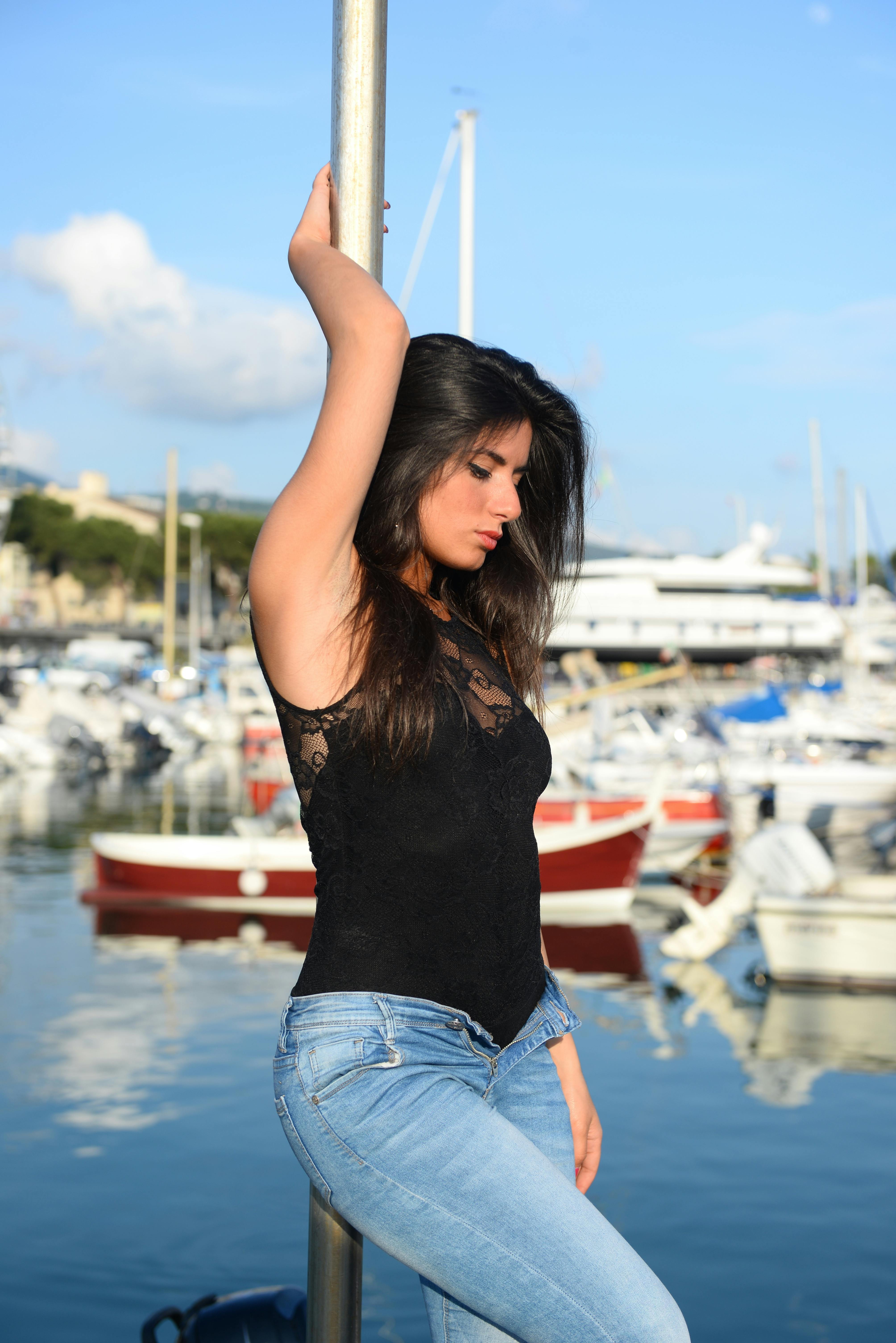 A fashionable woman poses by a marina, showcasing casual chic style in a sunlit coastal setting.
