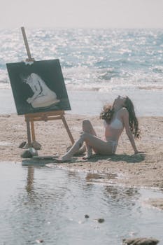 A woman in a bikini enjoys a tranquil beach setting while painting by the ocean.