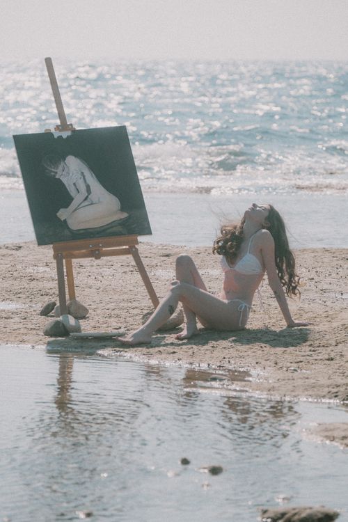 Free A woman in a bikini enjoys a tranquil beach setting while painting by the ocean. Stock Photo