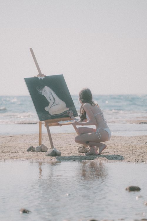 Free A woman painting on a beach easel captures the serene ocean view under sunlight. Stock Photo
