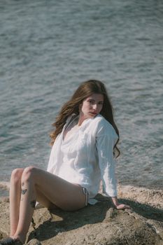 A woman in a white shirt sits on rocks by the sea, embracing natural beauty and tranquility.