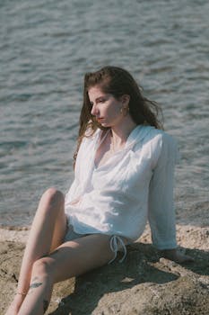 A serene portrait of a woman in a white outfit sitting on a rock by the ocean during summer.