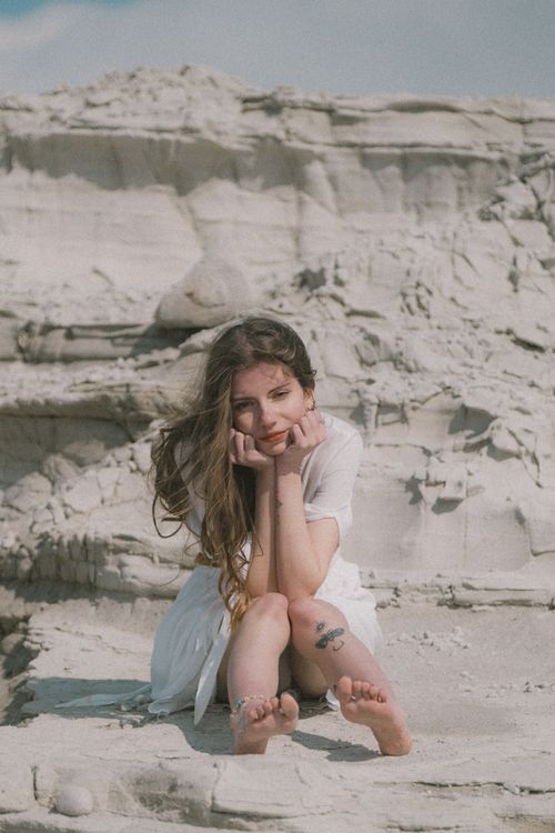 Free Artistic portrait of a woman in a bohemian style, sitting in a desert landscape during golden hour. Stock Photo