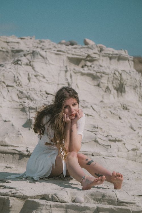 Free A bohemian woman in a white dress sits on rocks under natural light with a serene expression. Stock Photo
