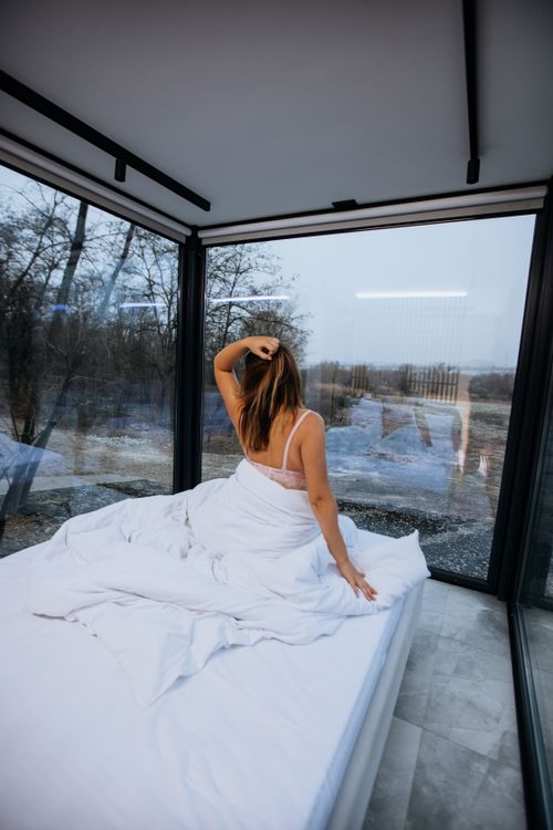 Free Woman sitting on bed, wrapped in white blanket, gazing at nature view through glass walls. Stock Photo
