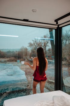 A woman in silk attire drinks coffee, gazing outside from a modern glass room.