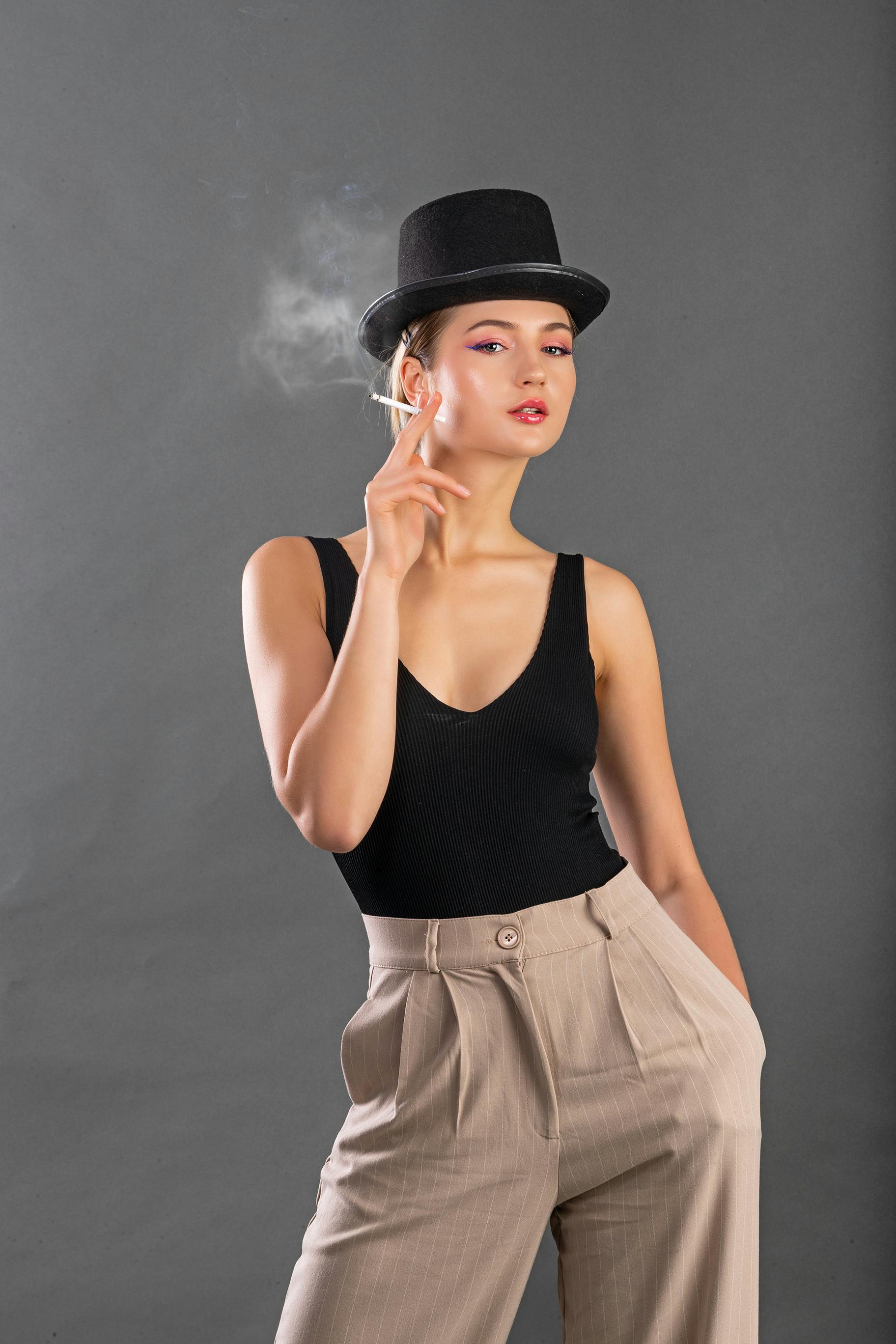 Gratis Model wanita bergaya mengenakan topi tinggi berpose dengan rokok di latar belakang abu-abu. Foto Stok
