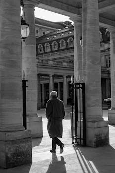 Black and white image of an elderly man walking among classic columns in a historic building.